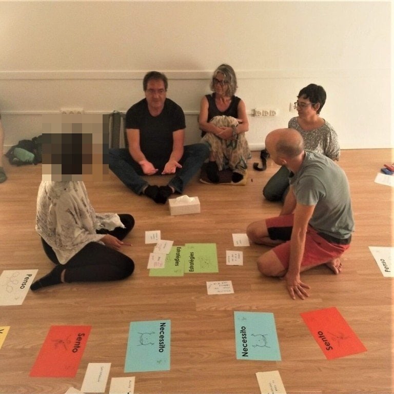Cinco personas sentadas en el suelo en círculo, rodeadas de grandes papeles de colores y tarjetas con palabras, participan en una actividad de grupo en el interior. El rostro de una persona aparece difuminado para preservar su intimidad.