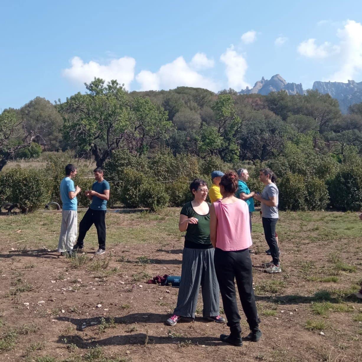 Varias personas permanecen en parejas al aire libre, en suelo seco, conversando o realizando actividades, con árboles y montañas de fondo bajo un cielo parcialmente nublado.
