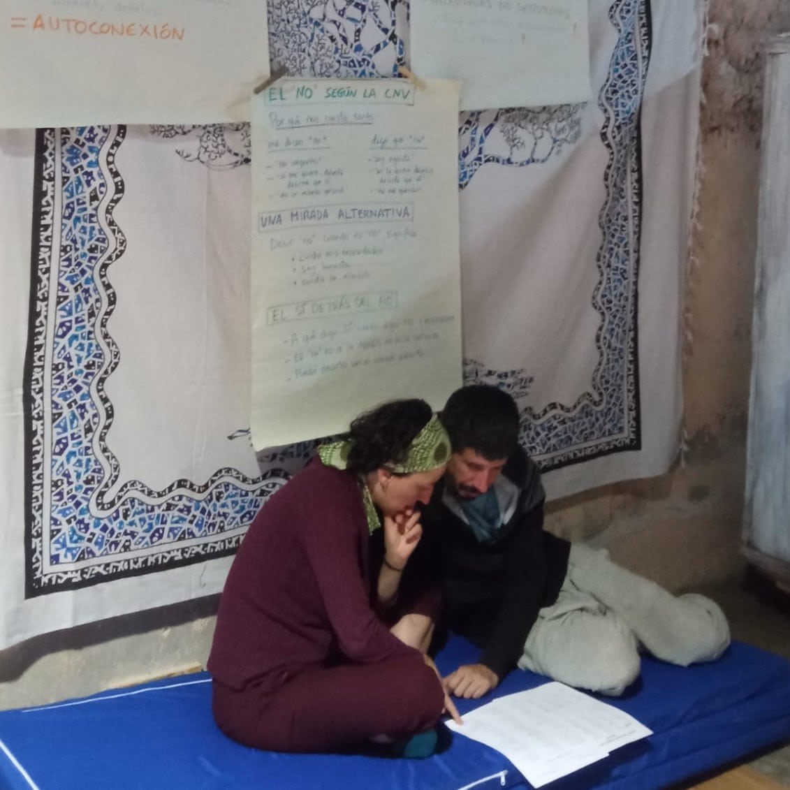 Dos personas sentadas en un colchón azul leen juntas un documento. Detrás de ellos, grandes hojas de papel con notas manuscritas están pegadas a una tela estampada en la pared.