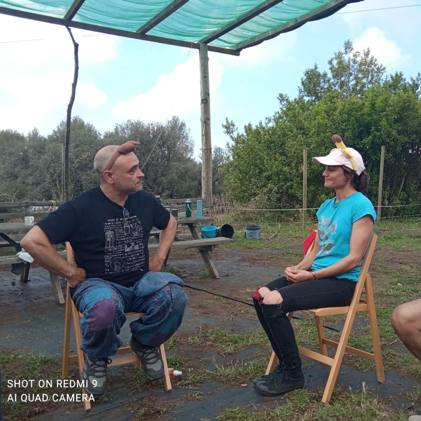 Dos personas sentadas en sillas frente a frente al aire libre, ambas con diademas con orejas falsas de animales. Al fondo hay una mesa de picnic de madera y vegetación.