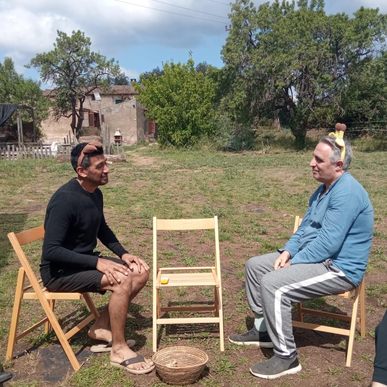 Dos hombres sentados en sillas de madera frente a frente al aire libre, con una cesta en el suelo y una silla vacía entre ellos. Uno de ellos lleva una cinta en la cabeza con un pequeño objeto.