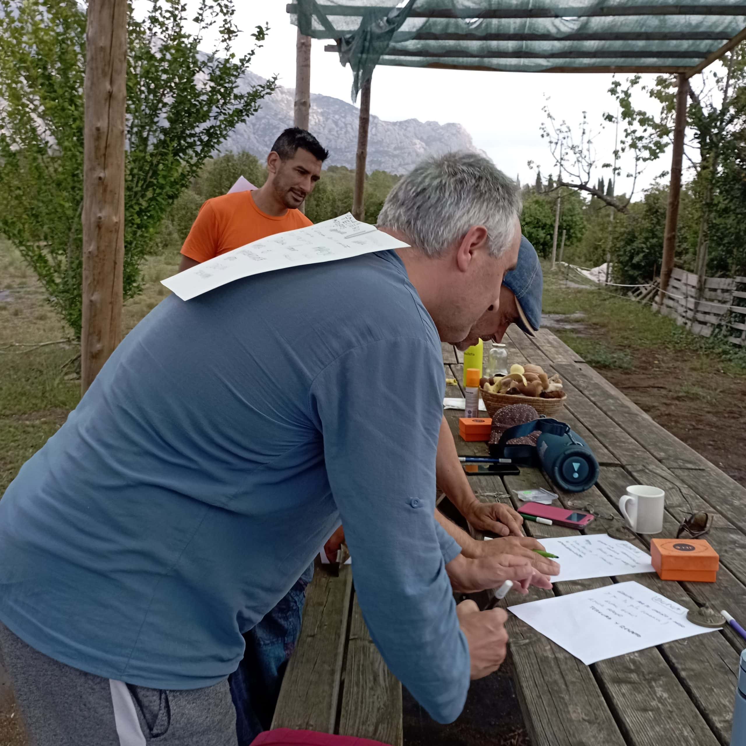 Tres personas trabajan en una mesa de madera al aire libre con papeles y artículos de papelería; una persona tiene una hoja de papel apoyada en la espalda. Al fondo se ven árboles y montañas.