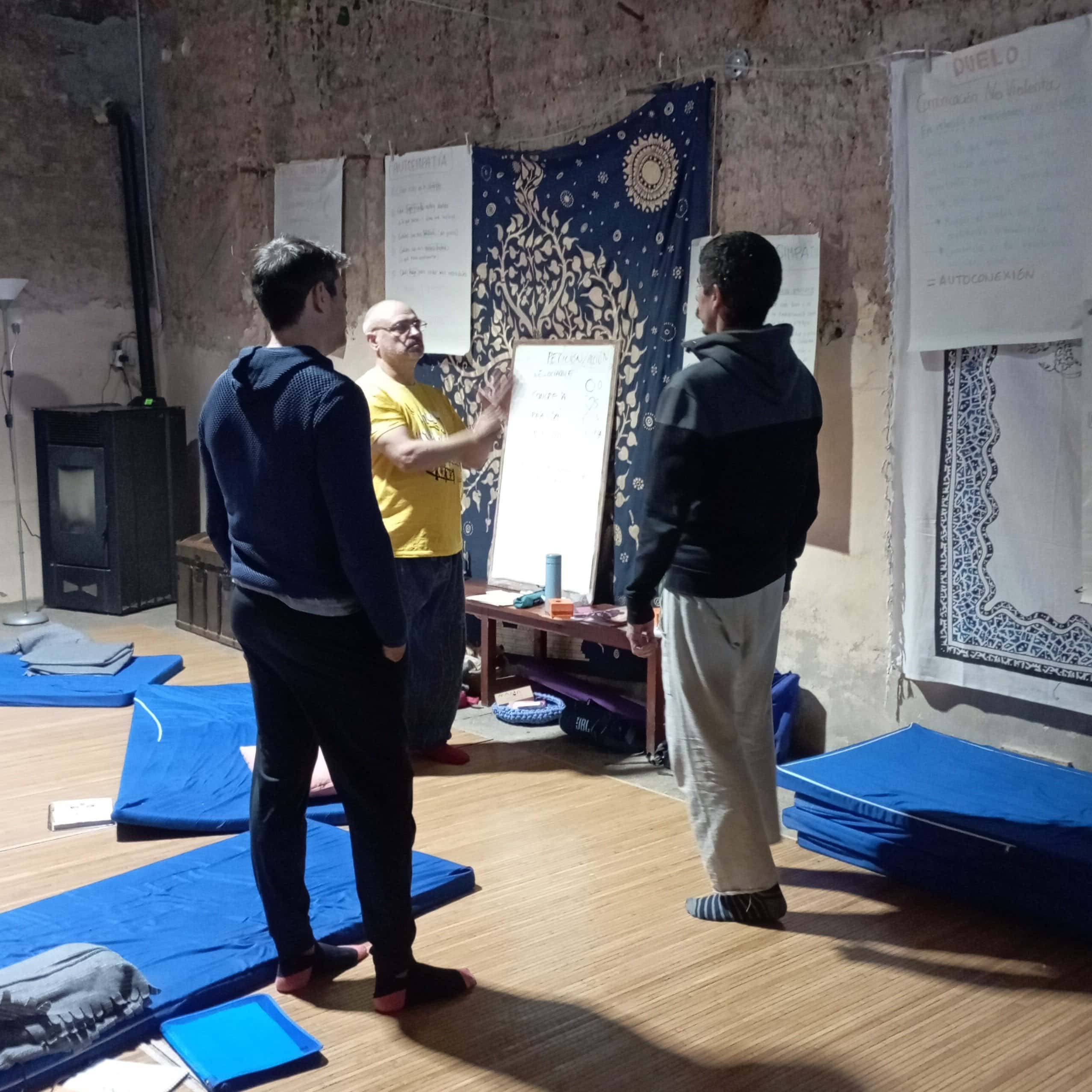 Tres personas están de pie en una habitación con alfombras azules en el suelo, manteniendo una discusión cerca de una pizarra blanca y grandes hojas de papel con notas en la pared.