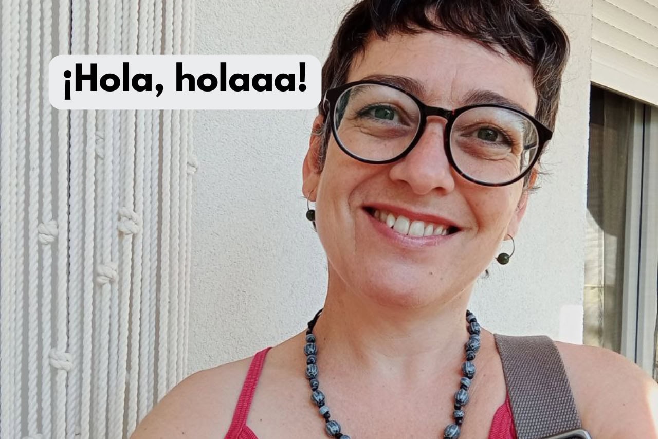Una persona con pelo corto y oscuro, gafas y un collar sonríe a la cámara. El fondo muestra una pared blanca y una cortina. El texto de la imagen dice: "¡Hola, holaaa!.