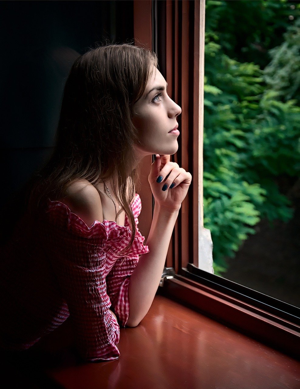 Una mujer con un top rojo a cuadros mira pensativa por una ventana abierta, con el follaje verde visible en el exterior.
