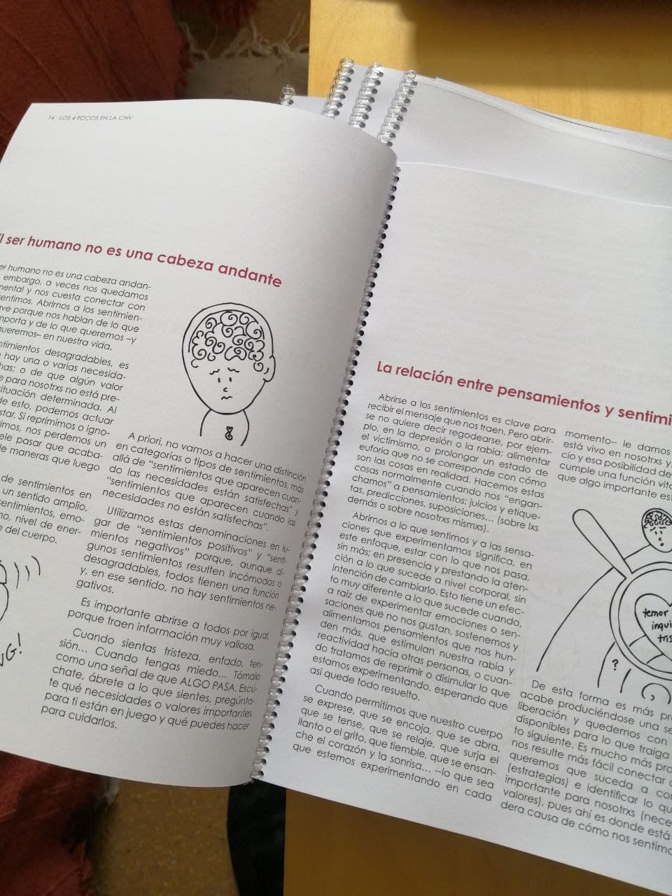 Sobre una mesa hay dos libros abiertos, encuadernados en espiral y en español, que muestran páginas con texto y sencillas ilustraciones en blanco y negro de cabezas y cerebros.