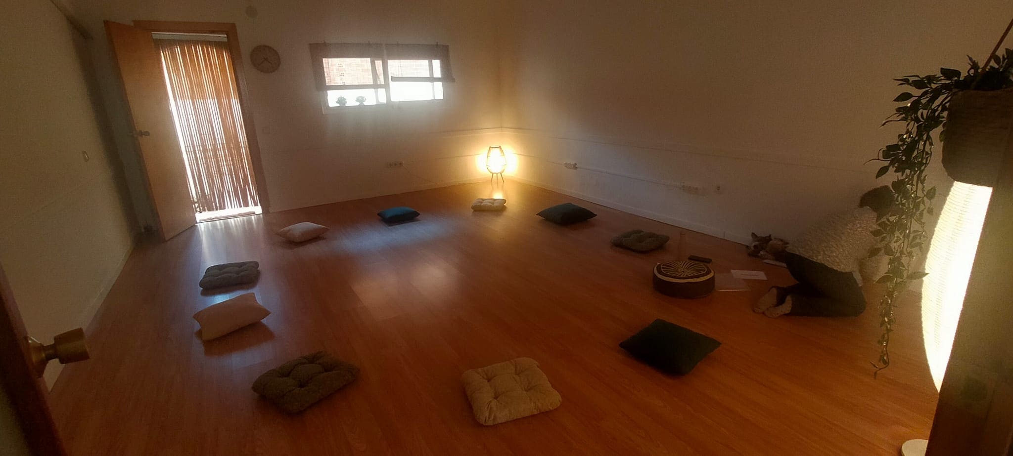 Una habitación escasamente amueblada con cojines dispuestos en círculo sobre un suelo de madera, iluminación tenue y una persona sentada cerca de la pared con papeles y un tambor.