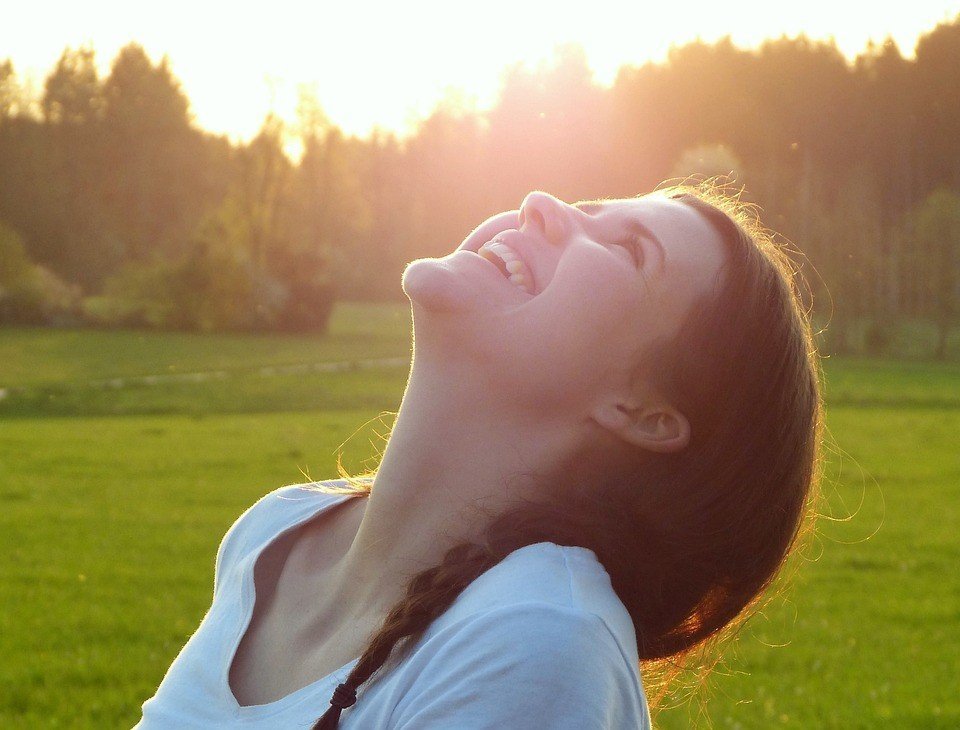 Una joven con una trenza se echa hacia atrás y sonríe al aire libre en un campo cubierto de hierba, con la luz del sol brillando tras ella: irradia alegría y confianza, inspirada por el coaching para materializar lo que quiero.