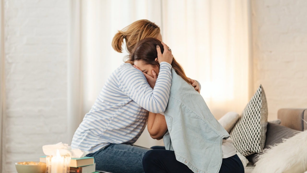 Dos mujeres sentadas se abrazan, una de ellas está llorando y la otra parece una amiga que la consuela