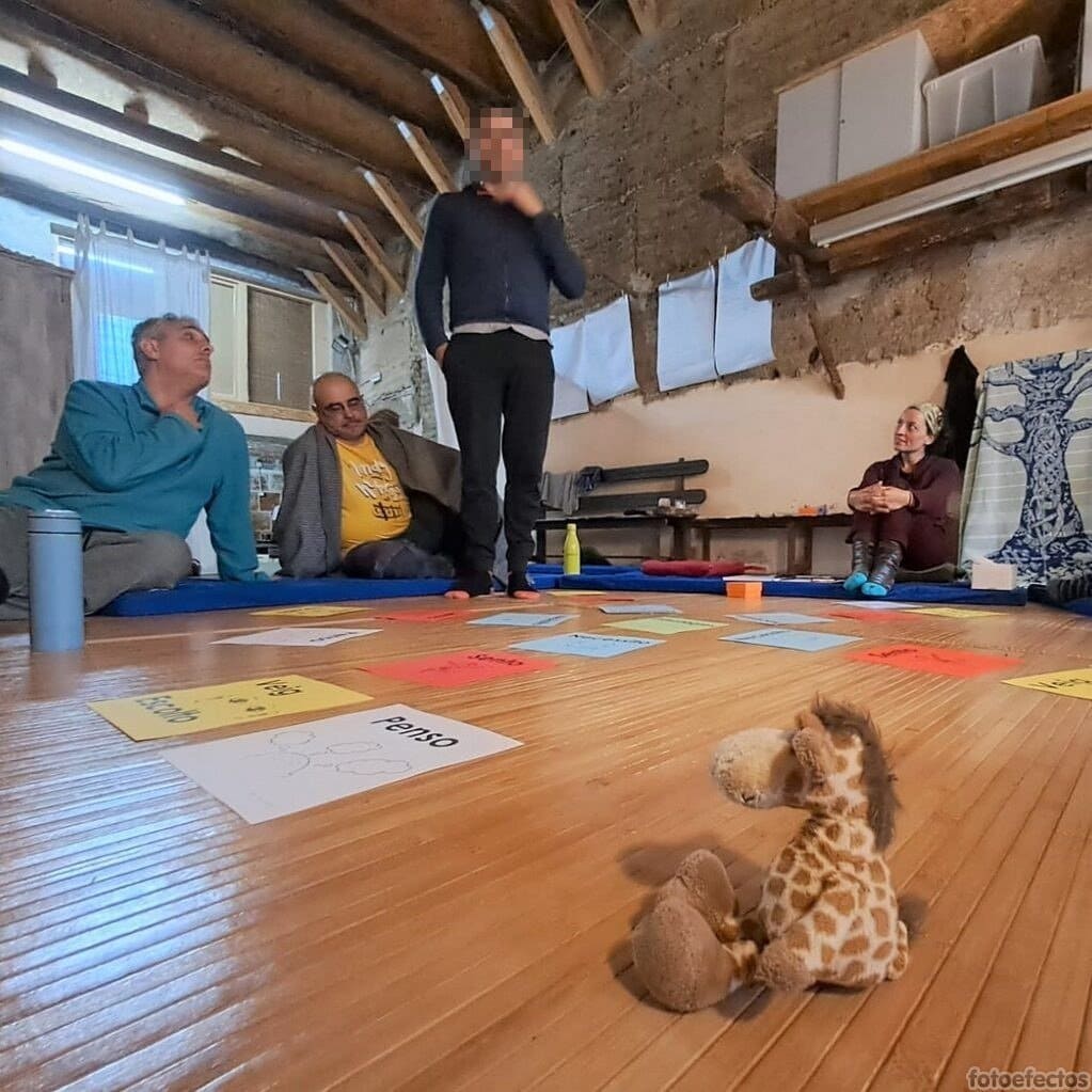 Cuatro personas están sentadas o de pie sobre colchonetas en una habitación rústica, mientras una jirafa de peluche descansa en el suelo de madera rodeada de tarjetas de papel.