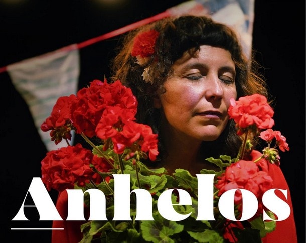 Una mujer con el pelo rizado y una flor roja en el cabello está de pie con los ojos cerrados, sosteniendo un ramo de flores rojas. En la parte inferior aparece la palabra "Anhelos".