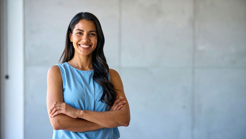 Mujer sonriendo con brazos cruzados en fondo neutro.