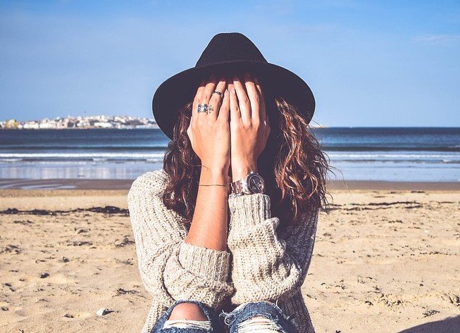 Una persona con sombrero y jersey está sentada en una playa de arena con las manos cubriéndose la cara, reflexionando en silencio; el océano y los edificios lejanos se ven al fondo, evocando un momento de comunicación noviolenta con uno mismo.