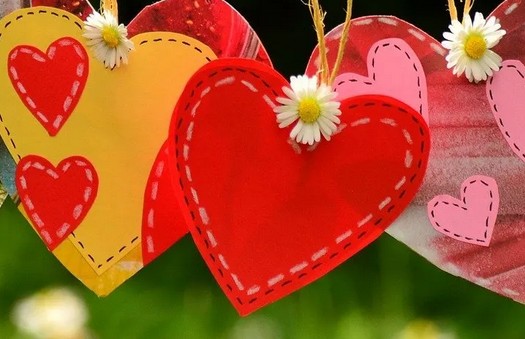 Corazones de papel en rojo, amarillo y rosa están decorados con pequeñas margaritas blancas y motivos cosidos, y cuelgan al aire libre sobre un fondo verde difuminado.