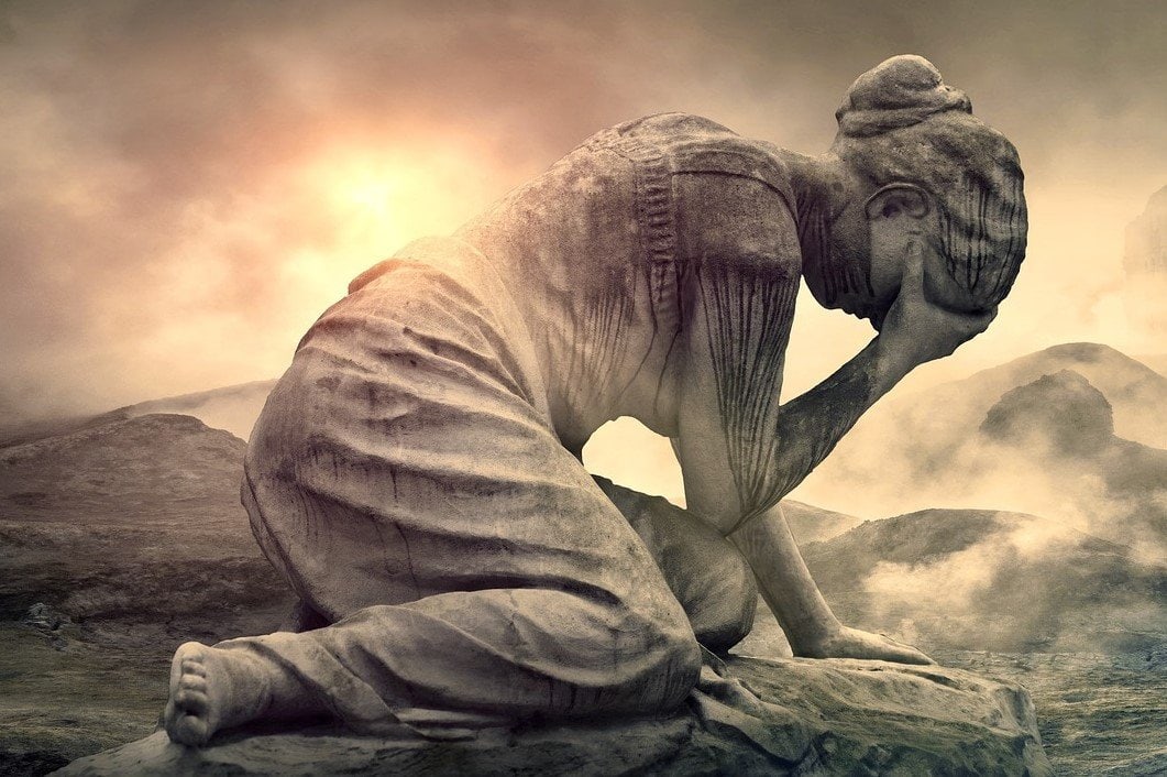 Una estatua de piedra erosionada de una mujer arrodillada, con la cabeza en la mano, sobre un paisaje brumoso y dramático en el que la luz del sol se abre paso entre las nubes.