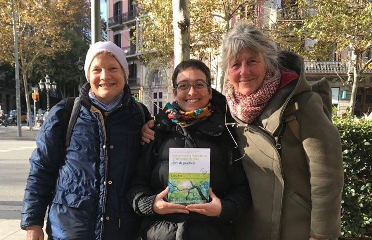 Tres mujeres están de pie al aire libre en una ciudad, sonriendo a la cámara. La mujer del centro sostiene un libro titulado "Libro de prácticas". Al fondo se ven árboles y edificios.
