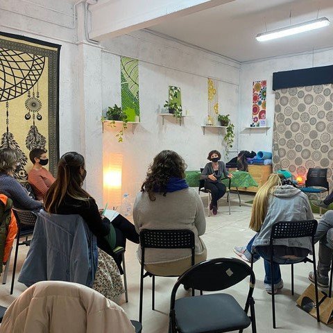 Un grupo de personas se sienta en semicírculo en sillas en el interior, escuchando a un orador situado en la parte delantera de la sala. Estanterías con plantas y tapices estampados decoran las paredes.