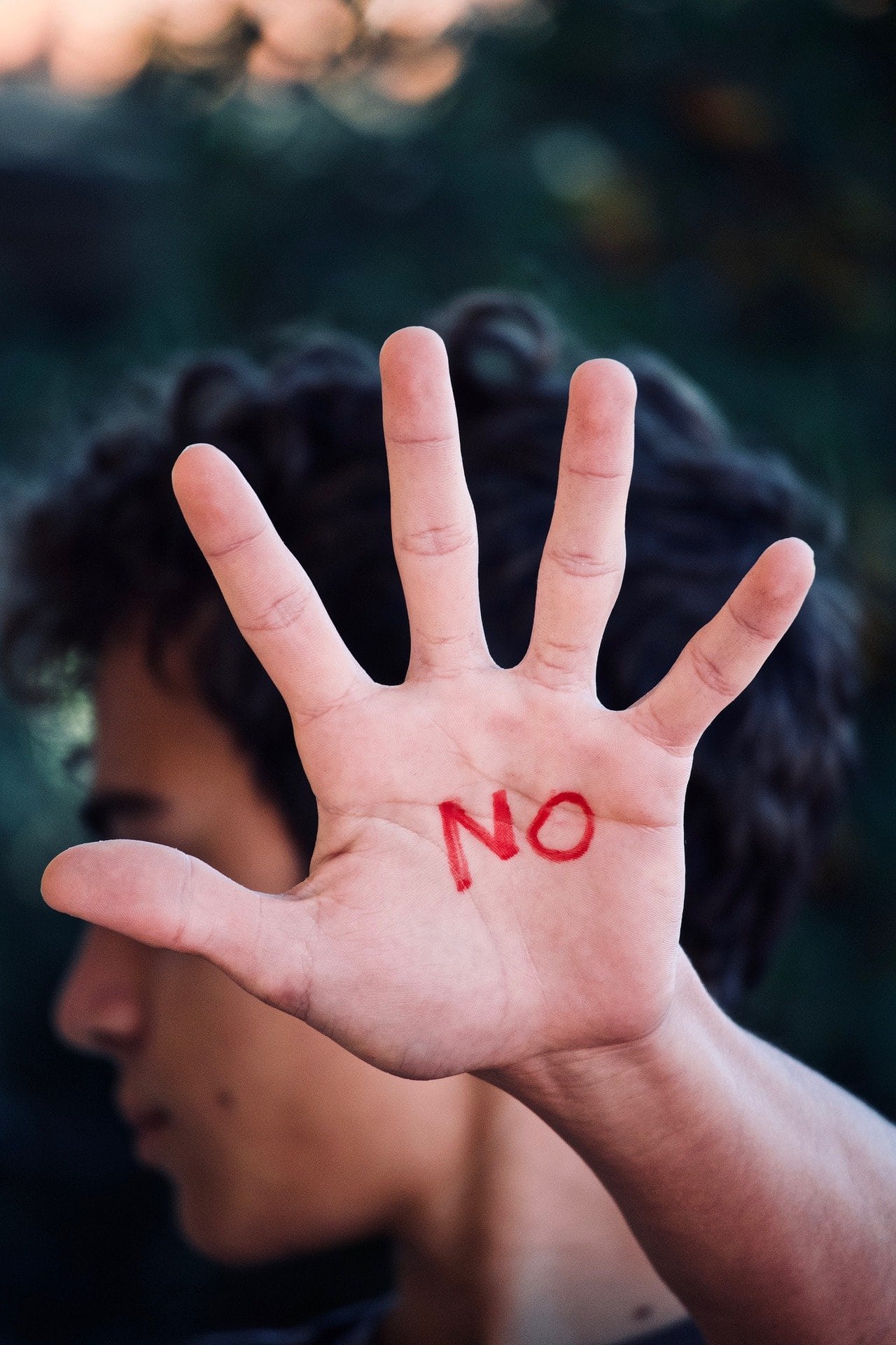 Una persona levanta una mano abierta con la palabra "NO" escrita en rojo en la palma, mirando a la cámara; su cara es parcialmente visible en el fondo.