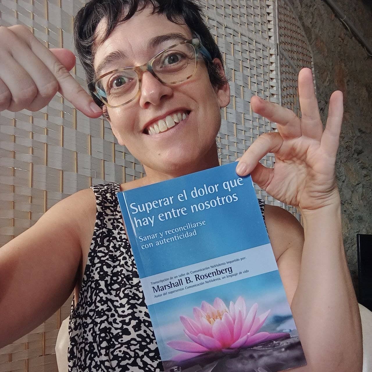 Una persona sonriente con gafas señala la portada de un libro en español titulado "Superar el dolor que hay entre nosotros", de Marshall B. Rosenberg.