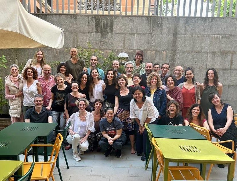 Un grupo de 32 adultos posan juntos al aire libre frente a un muro de piedra, con mesas verdes y sillas amarillas en primer plano.