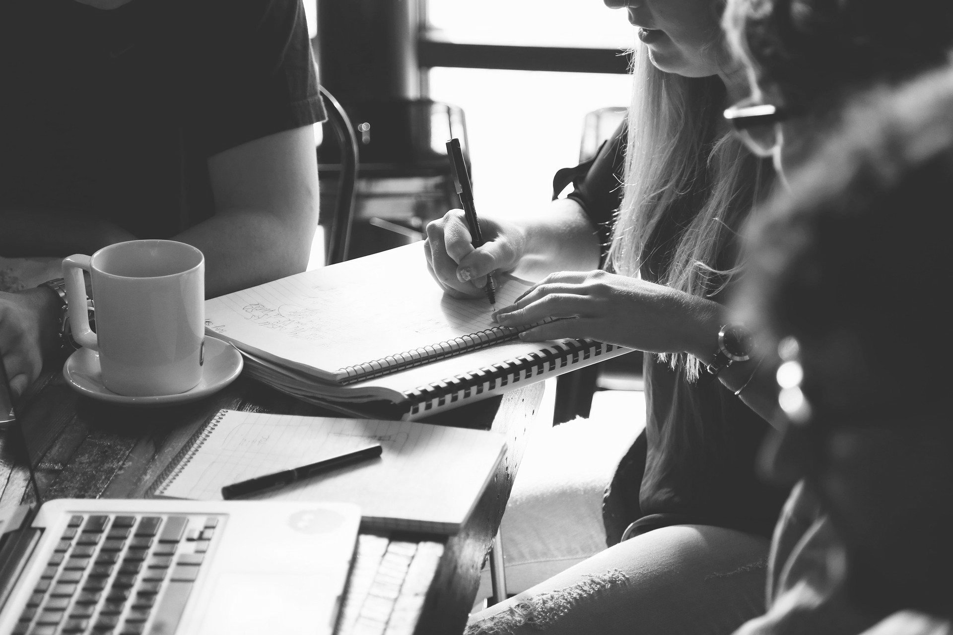 Grupo de personas sentadas a una mesa, tomando notas en cuadernos y utilizando un ordenador portátil, con una taza de café cerca.