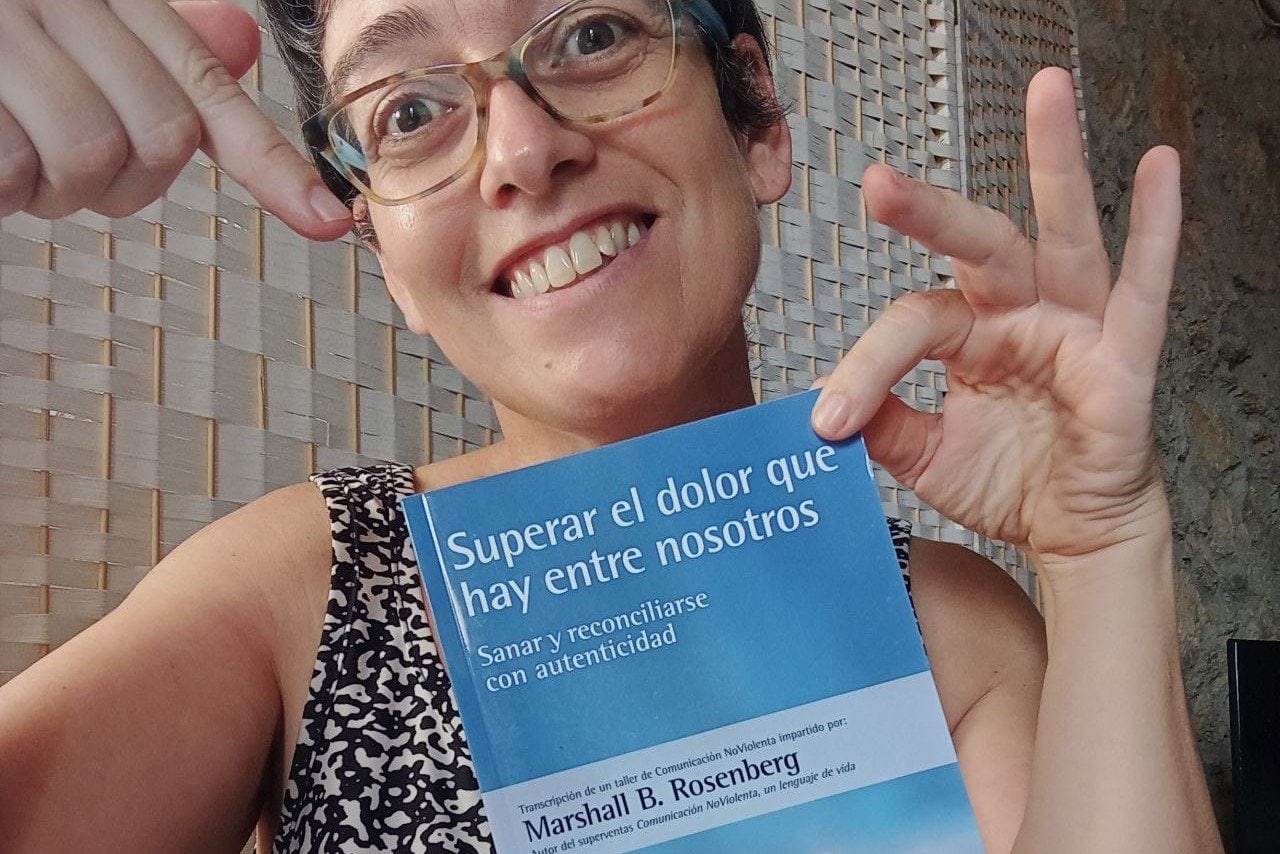 Una mujer con gafas sonríe y señala un libro titulado "Superar el dolor que hay entre nosotros", de Marshall B. Rosenberg.